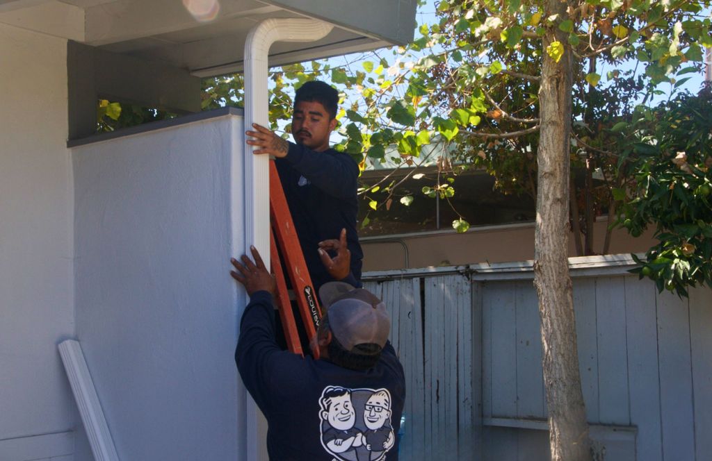 Two crew members on a ladder hanging a downspout under the eave